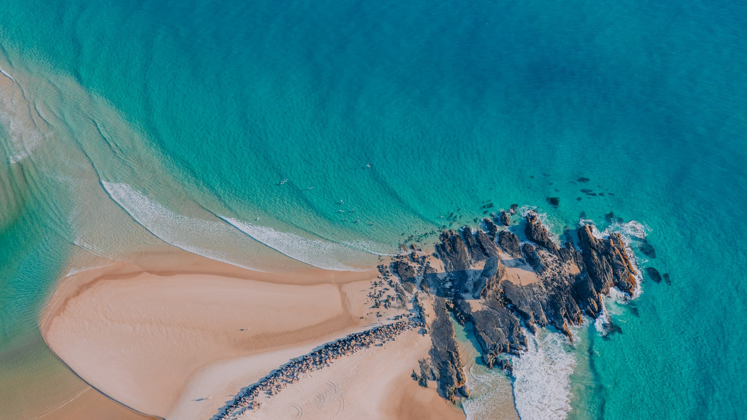 Aerial view of a bright turquoise coastline where surf meets a sweep of pale sand and dark rocks